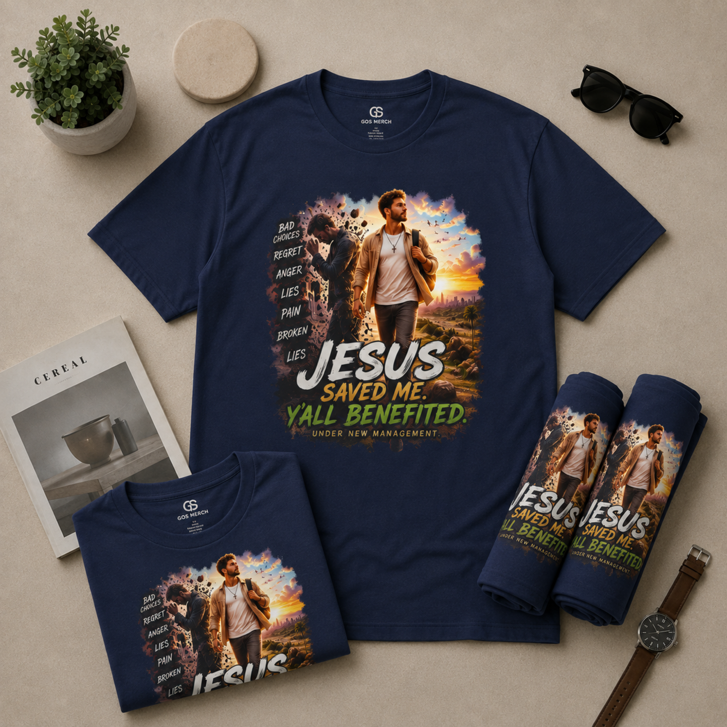 A navy blue graphic T-shirt laid flat on a beige surface, shown alongside a smaller folded version of the same shirt, rolled T-shirts, sunglasses, a plant, a watch, and a magazine. The main shirt features a large front print with a man walking outdoors and the text 'JESUS SAVED ME Y'all Benefited' and additional slogan text on the sleeves.