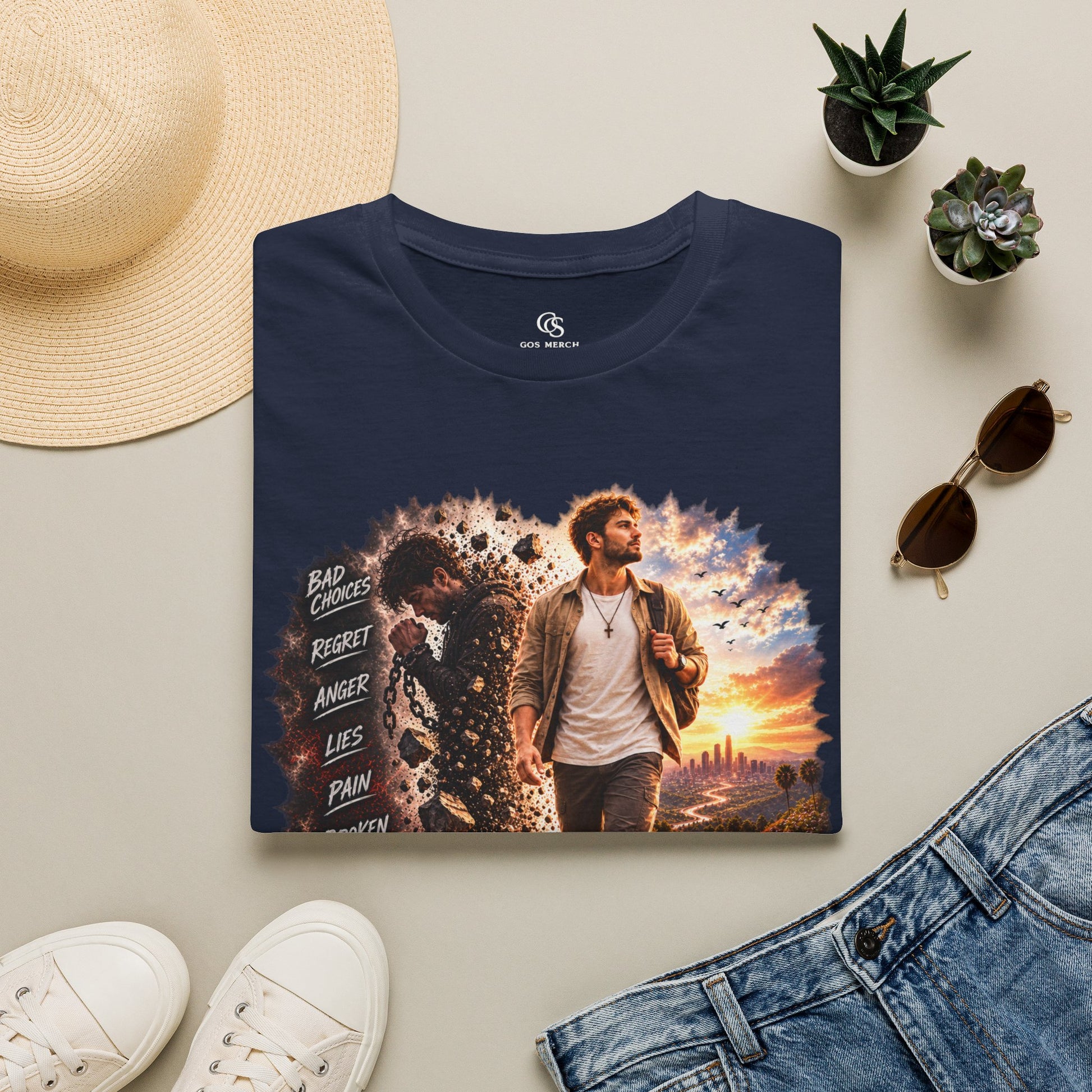 A navy blue graphic t-shirt featuring a printed design of a man walking forward with a dramatic sunset and text on the front, styled in a flat lay with a straw hat, white sneakers, denim shorts, and small potted succulents on a light beige background.