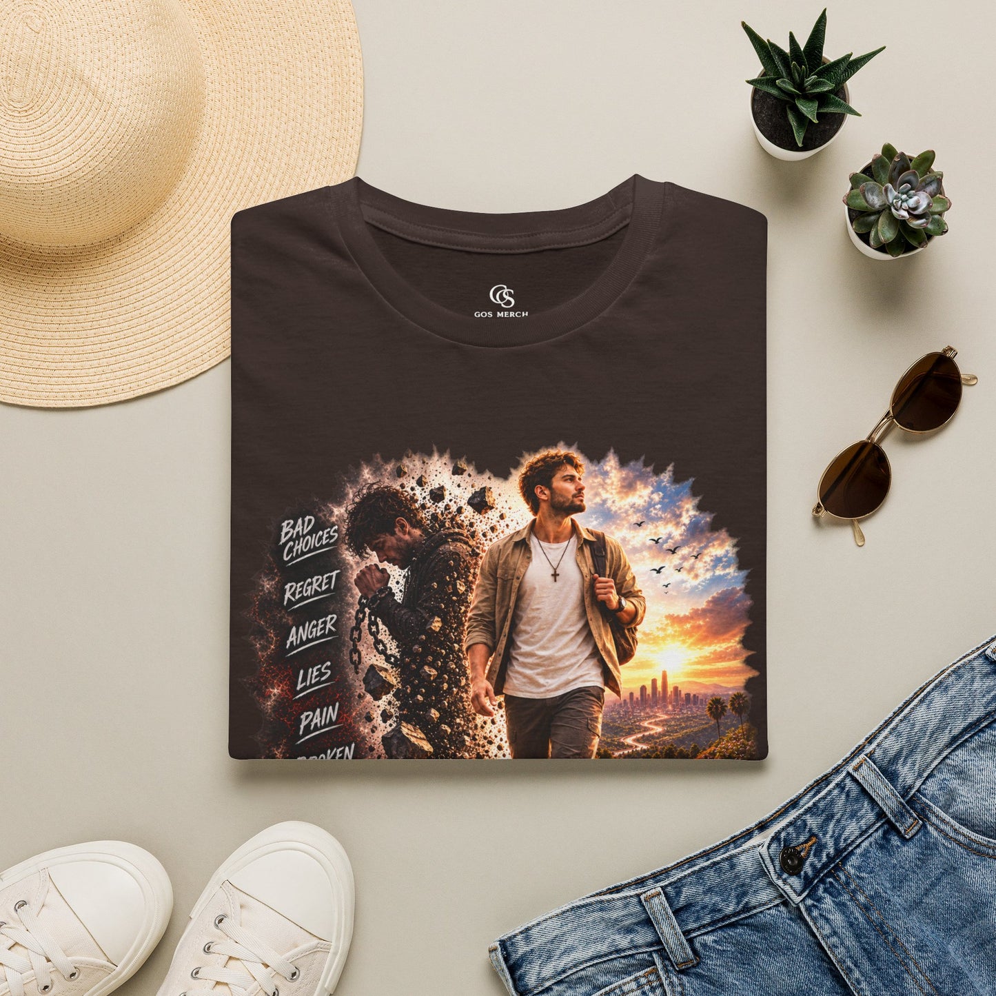 A dark brown t-shirt with a graphic print of a man walking forward against a dramatic sunset sky, surrounded by a styled flat-lay arrangement including a straw hat, white sneakers, sunglasses, and denim jeans on a light beige background.